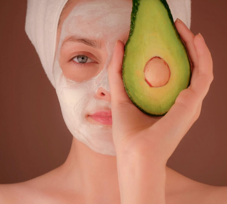 Woman with a white face mask holding half an avocado near her eye, promoting natural skincare.